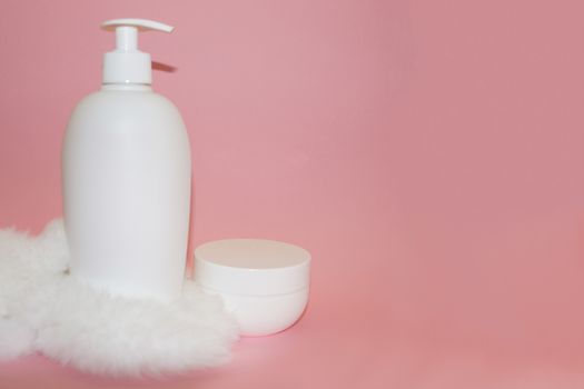 white cosmetic bottle, cream jar and white fur on pink background close-up