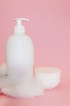 white cosmetic bottle, cream jar and white fur on pink background close-up