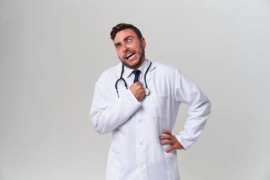 Handsome modern doctor white medical gown stands in studio gray background fool around Student medical university Positive medical worker Intern uniform looking at the camera funny grimaces choking