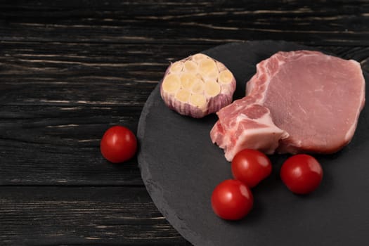 Top view of one pieces raw pork chop steaks with cherry tomatoes and garlic on a black stone cutting board.. Wooden table.