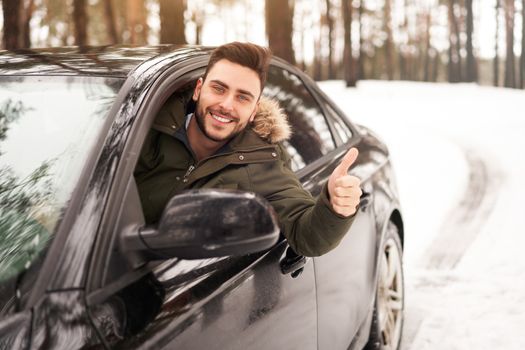 Young adult attractive Caucasian man sits at the wheel of his car sunny winter day shoeing thumbs up gesture. Wintertime road trip. Happy smiling hipster guy sitting in car and looking window.