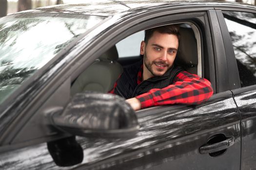 Young adult attractive Caucasian man sits at the wheel of his car sunny winter day. Wintertime road trip. Happy smiling hipster guy sitting in car and looking window. Portrait Positive driver