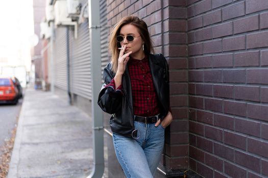 Beautiful young stylishly dressed Caucasian girl smokes cigarette on street Smoking bad habit Nicotine addiction Unhealthy lifestyle Modern woman leather jacket and jeans walking outdoor sunglasses