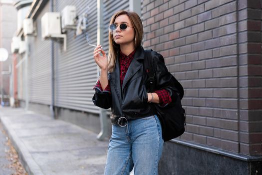 Beautiful young stylishly dressed Caucasian girl smokes cigarette on street Smoking bad habit Nicotine addiction Unhealthy lifestyle Modern woman leather jacket and jeans walking outdoor sunglasses