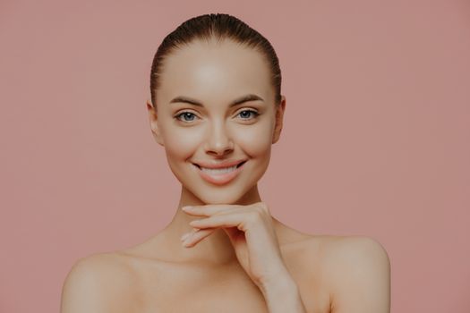 Pretty young European woman touches chin gently, enjoys flawless of skin after beauty procedures, poses nude, has natural makeup, isolated on pink background. Beauty, spa and wellness concept