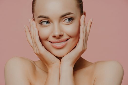 Face lift, skin care and wellness concept. Headshot of charming Caucasian young woman touches cheeks gently, has glowing hydrated skin, has bare soulders, looks tenderly aside. Beauty portrait.