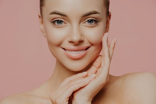 Close up shot of beautiful woman with naked shoulders, touches face gently, has healthy skin, natural makeup, looks directly at camera, isolated on pink background. Beauty, face care and spa concept