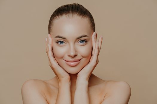 Close up portrait of blue eyed glad woman with dark combed hair, touches face gently, has healthy perfect skin, bare shoulders, enjoys beauty procedures, models indoor. Skin care and wellness