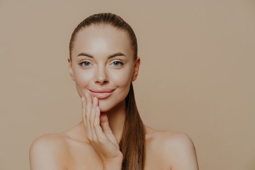 Close up portrait of beautiful naked young woman smiles tenderly and touches face, has healthy clean skin, cares about appearance, poses against beige background. Youth and skin care concept.