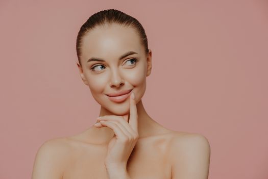 Topless thoughtful lady with clean perfect skin, dark hair, looks aside, undergoes facial trearrments, poses indoor, isolated over rosy background. Cosmetology, skin care and wellness concept