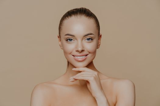 Cheerful young woman with clean glowing skin, keeps hand under chin, smiles gently, has well groomed body, combed hair, poses against beige background. Pure skin, beauty and wellbeing concept