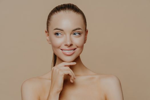 Smiling well groomed young woman with beauty face care, healthy glowing skin, touches chin gently and looks aside, poses shirtless against brown studio background, loves her skin, feels refreshed