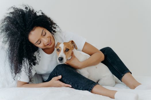Horizontal shot of lovely curly woman embraces favourite dog, wears whihte t shirt, jeans and socks, enjoy time together, sit on bed. Good beginning of day. Cheerfull lady plays with pet in bedroom