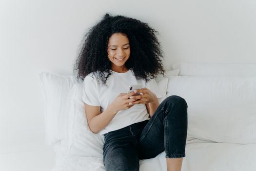 Relaxed beautiful Afro woman with cheerful expression uses modern mobile phone, surfs internet, poses in white comfortable bed, connected to wifi, browses webpage, enjoys domestic atmosphere