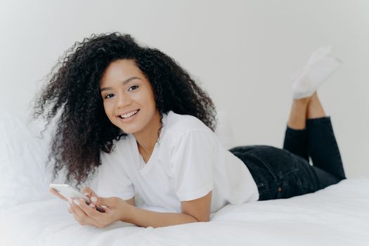 Horizontal shot of curly ethnic woman with pleasant smile, lies on comfortable bed, raises legs, uses modern cellphone, reads text message, enjoys high speed internet connection. People and bed time