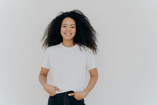 Isolated shot of young African American woman wears white t shirt, expresses good emotions, stands alone indoor, poses for photo, has casual talk with friend, enjoys free time. People and happiness