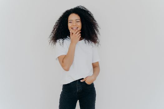 Overjoyed mixed race curly woman being in good mood, holds chin and giggles happily, keeps hand in pocket, dressed in casul wear, poses against white studio wall, has fun, hears pleasant news