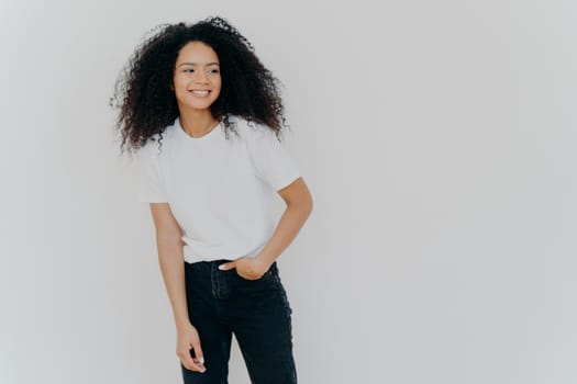 Young curly haired lady has slim figure, wears white t shirt and jeans, looks aside with happy expression, shows healthy teeth, notices funny scene on right side, isolated on white background