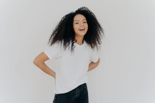 Portrait of cheerful millennial woman keeps hands behind back, dressed in casual wear, laughs from something fun, isolated over white studio background. African American lady expresses happiness