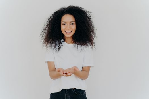 Glad dark skinned woman has cupped hands, asks to give something, smiles plesantly, has bushy frizzy hair, wears t shirt and jeans, isolated over white background. People and charity concept