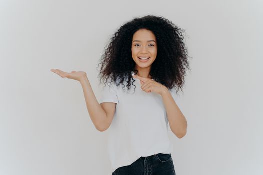 Choice is simple. Attractive feminine girl with Afro hair raises palm and points on blank space, makes decision, has toothy smile, wears casual wear, poses against white background. Look at this