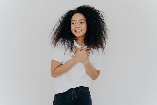 Photo of happy girl keeps both hands on chest, looks aside with pleasant smile, notices something awesome, poses against white background, wears casual t shirt and jeans. Positive emotions concept