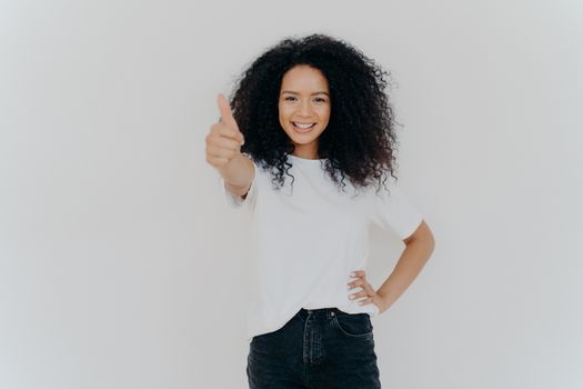 Photo of happy curly woman with crisp hair raises thumb up, gives approval, says sounds good, makes supportive gesture, keeps other hand on waist, dressed casually, isolated over white background