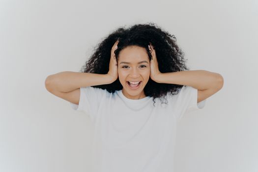 Photo of joyful dark skinned woman keeps hands on head, curly hair, laughs from joy, excited from positive emotions, dressed in white t shirt, hears funny joke, laughs sincerely. People, emotions