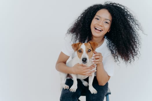 Portrait of joyful curly girl petting her dog, rejoicing buying jack russell terrier, smiles broadly, plays with animal, wears casual clothing, isolated over white background, enjoys good day