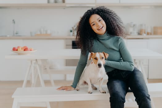 Female pet owner expresses love to dog, dressed in casual turtleneck and jeans, sits at white bench against cozy kitchen interior, spends free time at home, smiles broadly from happy enotions