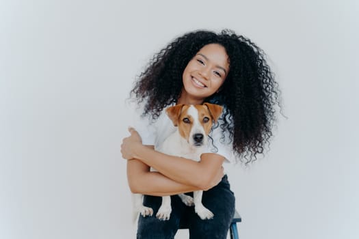 Horizontal shot of pleasant looking curly young woman embraces lovely pet, holds cute dog, smiles broadly, tilts head, isolated against white background. People, animals, good emotions concept