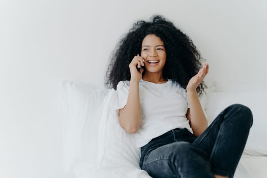 People, technology, communication and rest concept. Glad curly haired ethnic lady has telephone talk, gestures and laughs with joy, looks somewhere aside, spends time at home, sits on white bed