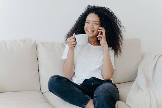 Cheerful Afro American woman has coffee break in living room, sits on couch, calls friend via modern gadget, has happy smile, thoughtful expression, discusses something pleasant. Technology, lifestyle