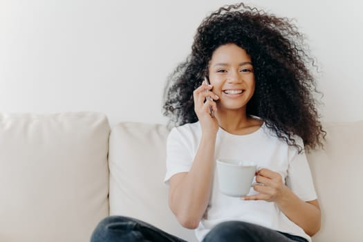 Horizontal shot of good looking woman has distant conversation via mobile phone, solves informal issues while talking, sits on sofa in living room, drinks tea, has broad smile, shows white teeth