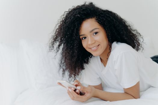 Shot of lovely curly woman with dark skin, tender smile on face, lies on white bed, wears t shirt, has relax with modern technologies, poses in bedroom, reads text message, uses free internet