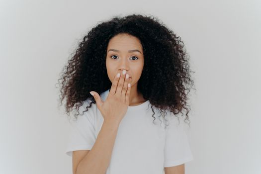 Headshot of impressed curly young woman covers mouth with palm, tries to be speechless, has curly hairstyle, manicure and minimal makeup, wears white t shirt, poses indoor. Omg, what gossip!