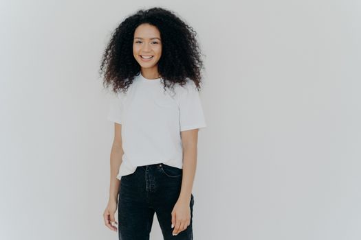 Picture of good looking woman with gentle smile, stands satisfied, has sincere carefree expression, relaxed casual mood, dressed in casual tshirt and black jeans, isolated over white background