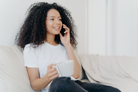 Sideways shot of positive ethnic brunette woman has telephone conversation, drinks hot beverage, holds white cup, sits on comfortable couch in living room, hears pleasant news, smiles happily