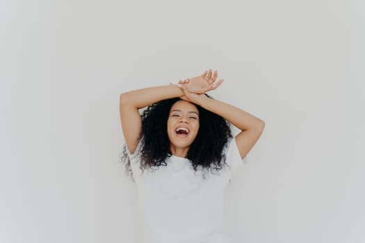 Overjoyed curly haired young lady keeps both hands on forehead, opens mouth and laughs happily, feels energetic, dressed in casual wear, poses against white studio background. What funny joke!