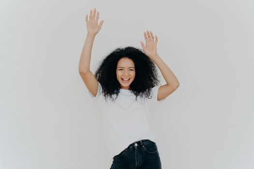 Half length shot of energetic carefree curly woman cheers with hands raised, wears white t shirt and jeans, has lucky day, perfect weekend, models against white background. Everybody put your hands up