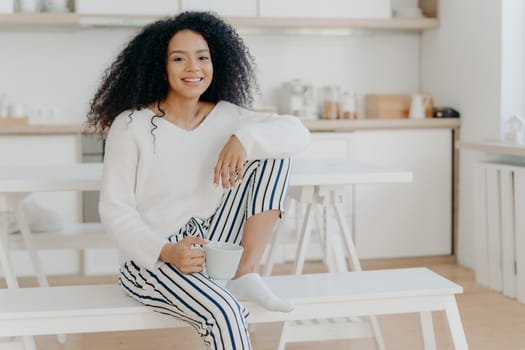 Glad African American woman holds cup of hot beverage, leans at knee, wears white stylish jumper and striped trousers, smiles pleasantly, spends leisure time at home, sits at bench in kitchen