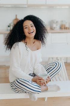 Vertical shot of pleased young Afro American woman feels relaxed and carefree, drinks aromatic coffee, sits crossed legs against kitchen interior, being in good mood, spends spare time at home