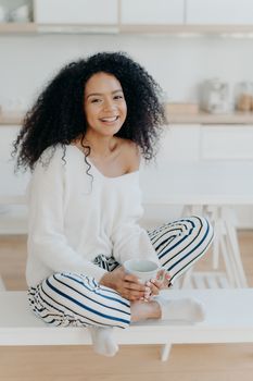 Image of lovely curly haired lady drinks coffee or tea from white mug, wears fashionable white sweater, striped pants, poses at kitchen against blurred background. People and lifestyle concept