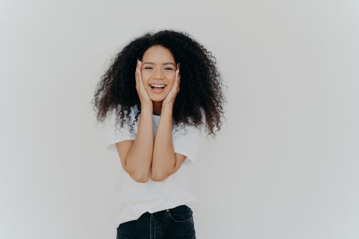 Candid shot of good looking lady with curly hairstyle, smiles cute, keeps both hands on cheeks, shows positive emotions, wears white t shirt, black denim trousers, stands indoor, blank copy space