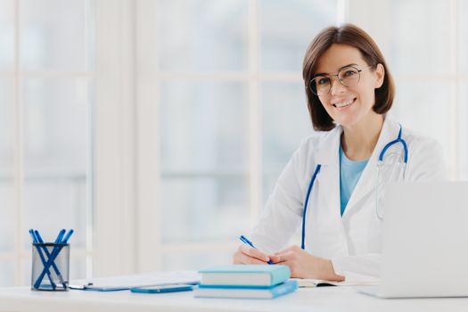 Professional woman doctor writes down notes, poses at desktop in office with laptop, wears white coat, spectacles and phonendoscope around neck, looks through medical documents. Healthcare concept