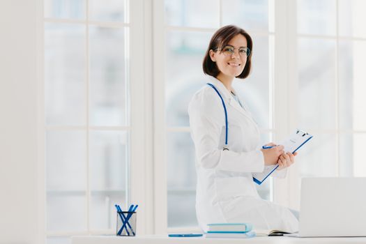 Skilled female physician writes in clipboard, poses at workplace with laptop computer. Professional family doctor ready to give you piece of advice to care about your health, wears medical uniform.