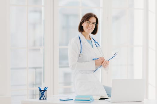 Skilled female therapist or cardiologist writes information at clipboard, ready to consult and cure patients, wears white coat, poses at hospital office with laptop computer, uses phonendoscope.