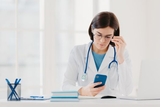 Medical technology concept. Concentrated woman doctor looks at smartphone device, checks necessary information in internet, wears stethoscope around neck, big round glasses, poses in clinic.