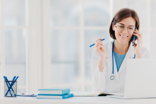 Photo of glad woman therapist or physician has phone conversation with patient, gives advice how to cure disease, wears white gown, reads information on laptop computer, works in private clinic