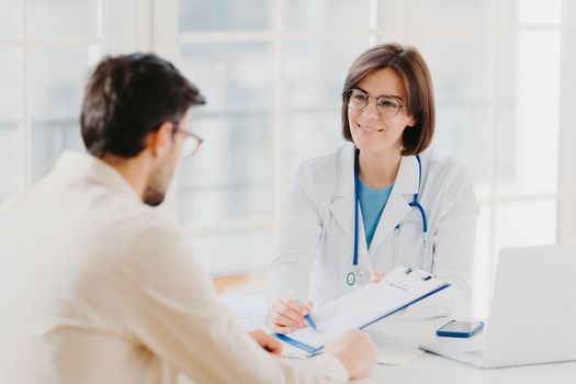 Young female doctor consults patient, shows information and prescription in clipboard, talk about medical checkup, pose at clinic, explains health insurance contract, read service agreement.